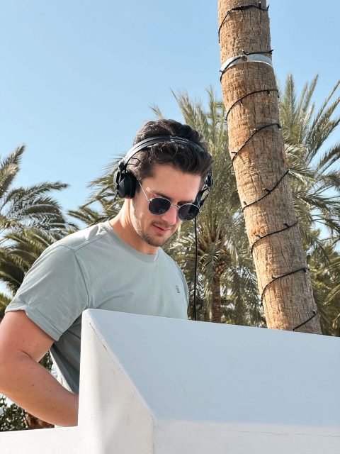 Hochzeitswahn - Ein Mann mit Sonnenbrille und Kopfhörern, möglicherweise DJ Fabian Baroud, steht draußen vor einem weißen Gebäude. Im Hintergrund sind Palmen und ein blauer Himmel mit Sonnenlicht zu sehen. Er blickt auf etwas vor ihm herab. Hochzeit in der Kulturfabrik Coburg.