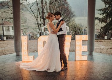 Hochzeitswahn - Braut und Bräutigam umarmen sich unter einem überdachten Bereich, hinter ihnen ein großes beleuchtetes „LOVE“-Schild. Die Braut trägt ein weißes Kleid, während der Bräutigam eine Weste und eine Krawatte trägt. Im Hintergrund sind Bäume und Hügel zu sehen. Zuckerbergschloss-Kappelrodeck-Hochzeit