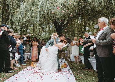 Hochzeitswahn - Braut und Bräutigam küssen sich, während der Bräutigam die Braut unter eine Weide taucht. Gäste umringen sie, klatschen und machen Fotos. Rosenblätter sind auf dem weißen Gang verstreut. Zur Dekoration gehören Laternen und weiße Luftballons. Hotel-Roessle-Hochzeit