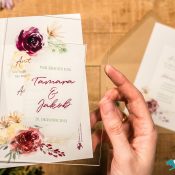 Hochzeitswahn - Zwei Hände halten eine Hochzeitseinladung mit Blumenmuster und dem Text „Art wir trauen uns“ und „Tamara & Jakob, 28. Oktober 2023“. Ein weiterer Umschlag und eine Einladungskarte, scheinbar automatisch erstellte Versionen, sind im Hintergrund auf einer Holzoberfläche zu sehen. Hochzeitseinladungen-selbst-gestalten