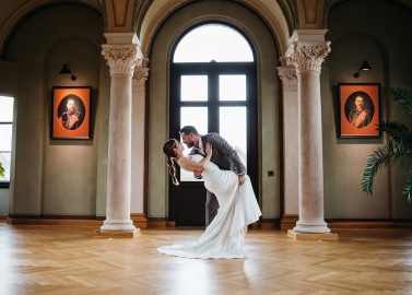 Hochzeitswahn - Ein Paar tanzt anmutig in einem eleganten Raum mit hohen Torbögen und dekorativen Säulen. Sie bewegen sich vor einem großen Fenster, flankiert von zwei Porträts. Der Holzboden glänzt unter ihren Füßen, während eine Topfpflanze der Szene Charme verleiht. Kaiserbahnhof-Halbe-Hochzeit