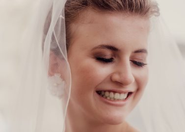 Hochzeitswahn - Eine Frau in Brautkleid und Schleier vor der ruhigen Kulisse des Vierwaldstättersees bei Luzern lächelt sanft mit geschlossenen Augen. Sie trägt elegante Ohrringe und eine Halskette, ihr Haar ist ordentlich gestylt. Das Bild strahlt einen weichen, warmen Ton aus und fängt die Essenz von „heiraten“ ein. Vierwaldstättersee-Schiffshochzeit