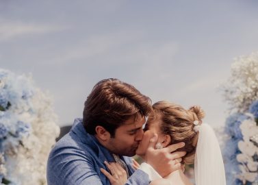 Hochzeitswahn - Ein Paar küsst sich während seiner Hochzeitszeremonie in der Nähe des Vierwaldstättersees. Der Bräutigam trägt einen blauen Anzug und die Braut ein weißes Kleid mit langem Schleier. Sie sind von Blumenarrangements mit blauen und weißen Blumen umgeben und feiern ihre Liebe in der bezaubernden Atmosphäre von Luzern. Vierwaldstättersee-Schiffshochzeit