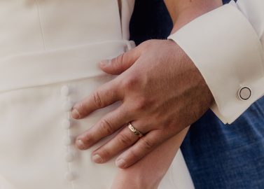 Hochzeitswahn - Nahaufnahme von zwei Personen, eine in einem weissen Outfit mit Knopfleiste und die andere in einem blauen Ärmel mit Manschettenknopf. Beide tragen Eheringe, eine Hand ruht über der anderen auf dem weissen Stoff – ein perfekter Moment, um inmitten des Charmes von Luzern in der Nähe des Vierwaldstättersees zu heiraten. Vierwaldstättersee-Schiffshochzeit
