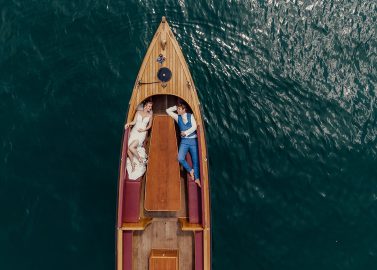 Hochzeitswahn - Luftaufnahme eines Holzboots auf dem ruhigen, dunkelgrünen Wasser des Vierwaldstättersees bei Luzern. Eine Frau in einem weißen Kleid und ein Mann in einem blauen Anzug liegen auf dem Deck und stehen sich gegenüber, als wären sie in einem zeitlosen Moment gefangen, was Gedanken ans Heiraten weckt. Vierwaldstättersee-Schiffshochzeit