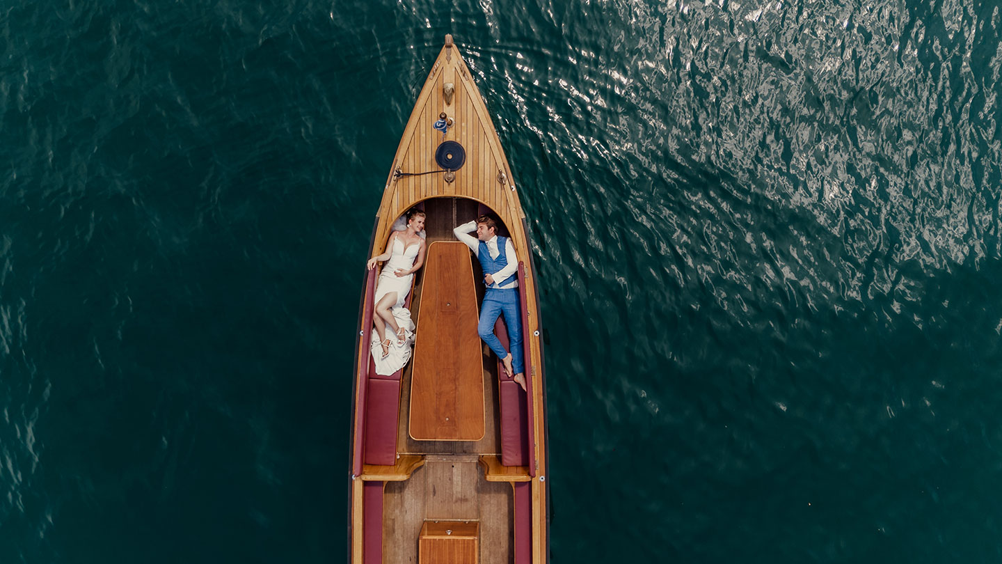 Hochzeitswahn - Luftaufnahme eines Holzboots auf dem ruhigen, dunkelgrünen Wasser des Vierwaldstättersees bei Luzern. Eine Frau in einem weißen Kleid und ein Mann in einem blauen Anzug liegen auf dem Deck und stehen sich gegenüber, als wären sie in einem zeitlosen Moment gefangen, was Gedanken ans Heiraten weckt. Vierwaldstättersee-Schiffshochzeit