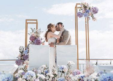 Hochzeitswahn - Ein Paar küsst sich vor einem Hochzeitsaltar im Freien, der mit violetten, rosa und weißen Blumen geschmückt ist. Dahinter stehen zwei geometrische Goldrahmen vor dem Hintergrund eines klaren blauen Himmels, was an die Eleganz des Hotels Rössle erinnert. Hotel-Roessle-Hochzeit