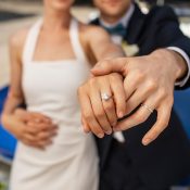 Hochzeitswahn - Ein Paar in formeller Kleidung hält den perfekten Moment fest, indem es seine Trauzeugen vor einem blauen Auto präsentiert. Die Frau präsentiert in einem weißen Kleid elegant ihren Verlobungsring und der Mann steht neben ihr und zeigt stolz seinen Ehering. Zuckerbergschloss-Kappelrodeck-Hochzeit