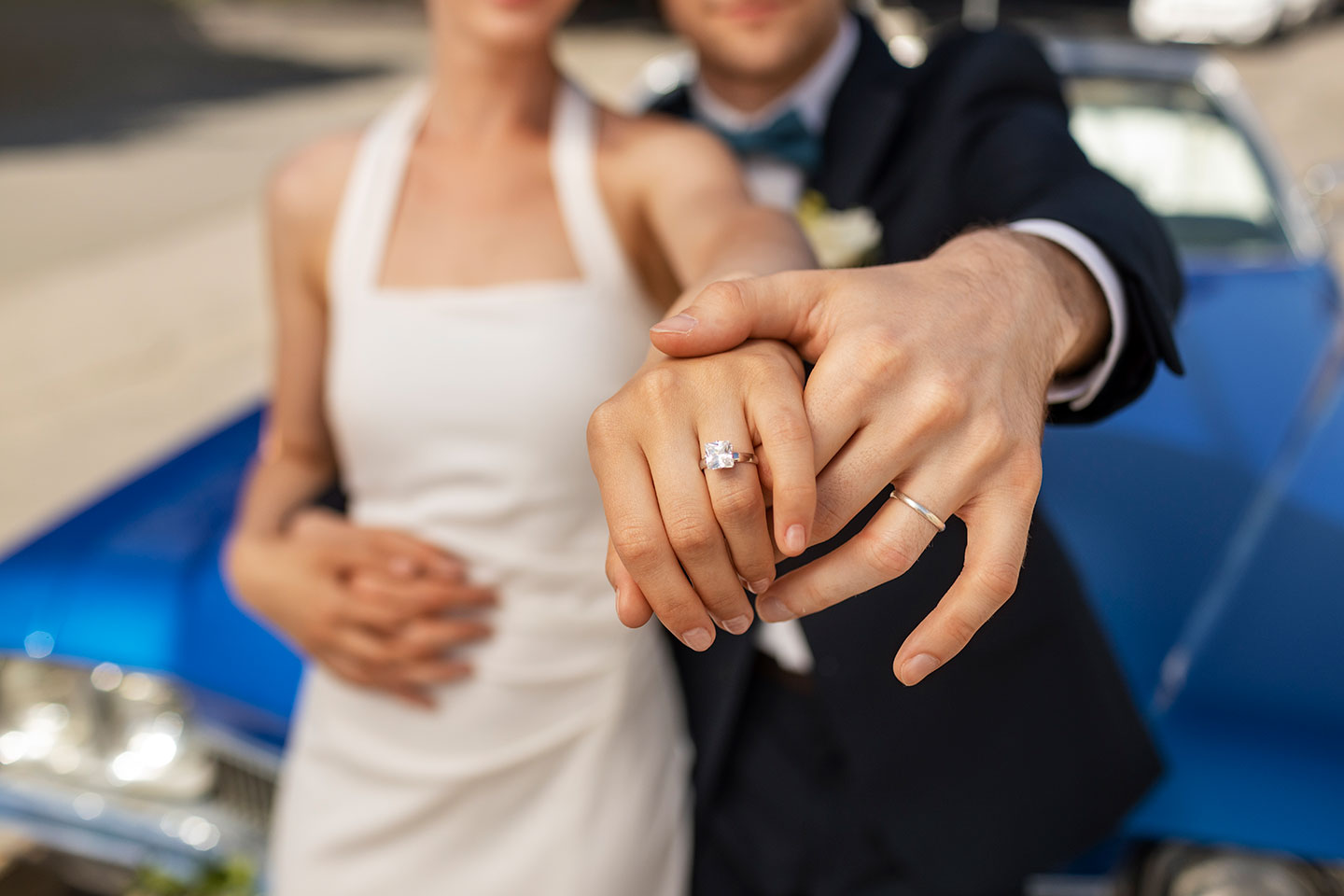 Hochzeitswahn - Ein Paar in formeller Kleidung hält den perfekten Moment fest, indem es seine Trauzeugen vor einem blauen Auto präsentiert. Die Frau präsentiert in einem weißen Kleid elegant ihren Verlobungsring und der Mann steht neben ihr und zeigt stolz seinen Ehering. Zuckerbergschloss-Kappelrodeck-Hochzeit