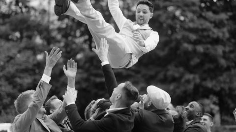 Hochzeitswahn - Eine Gruppe von Menschen, hauptsächlich Männer in Anzügen, wirft einen Mann in heller Abendkleidung in die Luft. Der Mann scheint zu lächeln oder zu lachen. Im Hintergrund sind Bäume und ein Team von McKenzie Productions zu sehen. Zuckerbergschloss-Kappelrodeck-Hochzeit