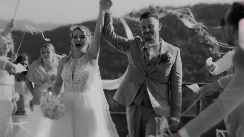 Hochzeitswahn - Braut und Bräutigam heben feierlich die Hände, während sie im Freien, umgeben von Gästen, den Gang entlanggehen. Diese von McKenzie Productions aufgenommene Schwarz-Weiß-Szene unterstreicht die Freude des Augenblicks auf wunderschöne Weise. Zuckerbergschloss-Kappelrodeck-Hochzeit