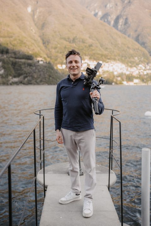 Hochzeitswahn - Ein Mann steht auf einem schmalen Steg über einem See und hält eine Kamera mit Stativ auf der Schulter. Er trägt ein dunkelblaues Langarmhemd, eine helle Hose und weiße Turnschuhe. Hinter ihm erheben sich Hügel und Gebäude – ein heiterer Moment, eingefangen von McKenzie Productions. Zuckerbergschloss-Kappelrodeck-Hochzeit