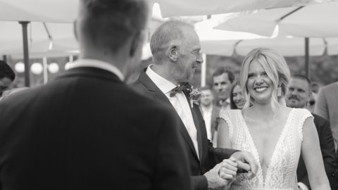 Hochzeitswahn - Eine Braut in einem weißen Kleid lächelt, als sie von einem älteren Mann im Anzug zum Altar geführt wird; im Hintergrund dieser Veranstaltung im Freien sind Menschen und Regenschirme zu sehen, die von McKenzie Productions wunderschön eingefangen wurden. Zuckerbergschloss-Kappelrodeck-Hochzeit