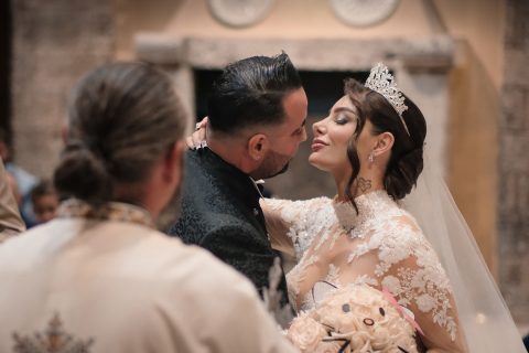Hochzeitswahn - Eine Braut in weißem Spitzenkleid und Tiara beugt sich vor, um den Bräutigam im dunklen Anzug zu küssen. Aufgenommen von Francesco Calarota Photography, halten sie einen Blumenstrauß in der Hand, während ein Geistlicher mit Pferdeschwanz in dieser eleganten Inneneinrichtung zusieht. Zuckerbergschloss-Kappelrodeck-Hochzeit