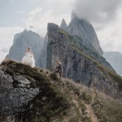 Hochzeitswahn - Eine Braut im weißen Kleid und ein Bräutigam im Anzug stehen auf einem grasbewachsenen, felsigen Berghang mit dramatischen, zerklüfteten Gipfeln und Wolken im Hintergrund, wunderschön eingefangen von Francesco Calarota Photography. Zuckerbergschloss-Kappelrodeck-Hochzeit