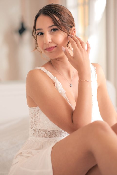Hochzeitswahn - Eine junge Frau mit braunem Haar und einem weißen Spitzenkleid sitzt auf einem Bett und berührt mit einer Hand ihr Gesicht. Mit einem leichten Lächeln blickt sie in die Kamera. Der sanft unscharfe Hintergrund unterstreicht die für FC Art von Francesco Calarota Photography typische Eleganz. Zuckerbergschloss-Kappelrodeck-Hochzeit