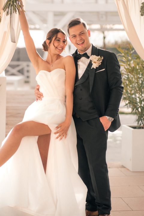 Hochzeitswahn - Eine Braut in einem weißen Kleid mit hohem Schlitz steht neben ihrem Bräutigam im schwarzen Smoking. Aufgenommen von Francesco Calarota Photography wirken beide glücklich – sie lächelt mit erhobenem Arm, während er sanft ihre Taille hält, während sie gemeinsam im Freien posieren. Zuckerbergschloss-Kappelrodeck-Hochzeit