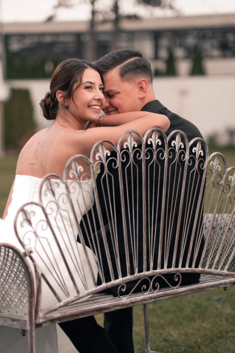 Hochzeitswahn - Eine Braut im weißen Kleid und ein Bräutigam im schwarzen Anzug sitzen eng beieinander auf einer dekorativen Metallbank im Freien. Aufgenommen von Francesco Calarota Photography, lächelt die Braut in die Kamera, während der Bräutigam sich vorbeugt und sie umarmt. Zuckerbergschloss-Kappelrodeck-Hochzeit