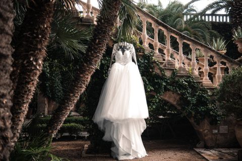 Hochzeitswahn - Ein weißes Brautkleid aus Spitze und Tüll hängt von einem Ast vor einer Steintreppe, umgeben von Grün und Palmen – eine bezaubernde Gartenszene im Freien, eingefangen von Francesco Calarota Photography. Zuckerbergschloss-Kappelrodeck-Hochzeit