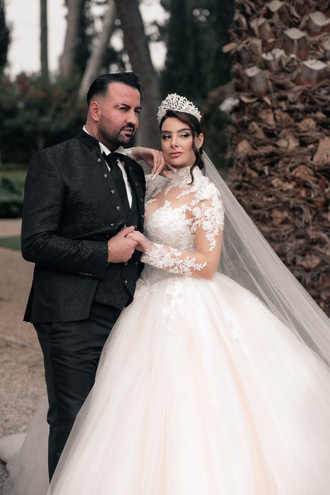Hochzeitswahn - Eine Braut in einem weißen Spitzenbrautkleid und einer Tiara steht neben einem Bräutigam im schwarzen Anzug. Sie halten Händchen und posieren im Freien neben einem grünen Baum. Ein wunderschönes Foto von Francesco Calarota Photography. Zuckerbergschloss-Kappelrodeck-Hochzeit