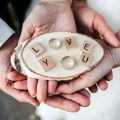 Hochzeitswahn - Hände halten eine Holzscheibe mit Scrabble-Steinen, auf denen „LOVE U“ steht. Gekrönt wird dies von einem Damenring und zwei weiteren Ringen, die die Buchstaben umschließen. Der Hintergrund ist unscharf und fängt einen flüchtigen Moment der Zuneigung ein. Zuckerbergschloss-Kappelrodeck-Hochzeit
