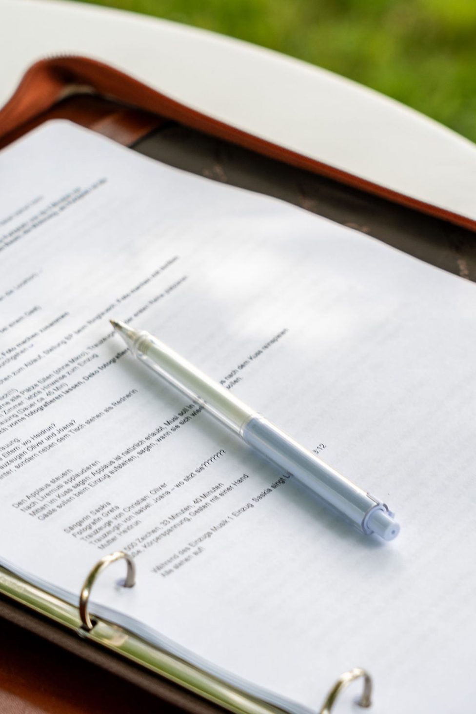 Hochzeitswahn - Ein Dreiringordner liegt auf einem Tisch und enthält bedruckte, mit Text übersäte Seiten. Ein transparenter Stift liegt diagonal über den Seiten. Der Hintergrund ist mit einem Hauch von Gras versehen und suggeriert eine Outdoor-Kulisse – perfekt für die Planung persönlicher Zeremonien und um dem neuesten Hochzeitstrend zu folgen. Zuckerbergschloss-Kappelrodeck-Hochzeit