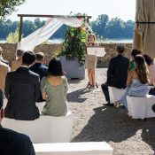Hochzeitswahn - Eine Frau steht an einem Rednerpult unter einem Holzbogen mit transparentem weißen Stoff und spricht zu den Gästen einer intimen Hochzeit mit Blick auf ein Gewässer. Die Versammlung, vor einer Kulisse aus Bäumen, präsentiert den neuesten Hochzeitstrend. Die Gäste sitzen in festlicher Kleidung auf weißen Bänken. Zuckerbergschloss-Kappelrodeck-Hochzeit