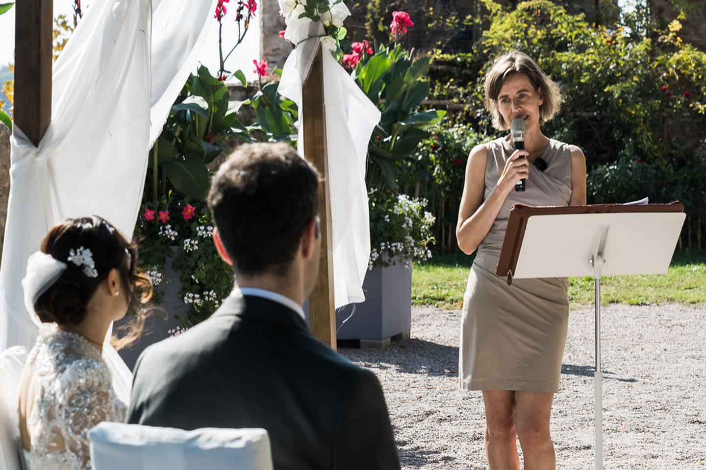 Hochzeitswahn - Eine Frau steht an einem Podium und spricht im Freien in ein Mikrofon zu einem sitzenden Brautpaar. Das Grün und die weißen Vorhänge unterstreichen den Charme der Hochzeitszeremonie und spiegeln den neuesten Hochzeitstrend für unvergessliche, persönliche Zeremonien wider. Zuckerbergschloss-Kappelrodeck-Hochzeit