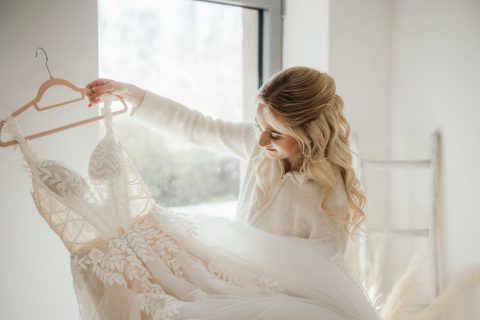 Hochzeitswahn - Eine Frau mit langen blonden Haaren hält ein weißes Brautkleid mit Spitzendetails und transparenten Einsätzen hoch und bewundert es am Fenster – ein Moment der Freude während ihrer Hochzeitsplanung mit Nelyce Weddings.
