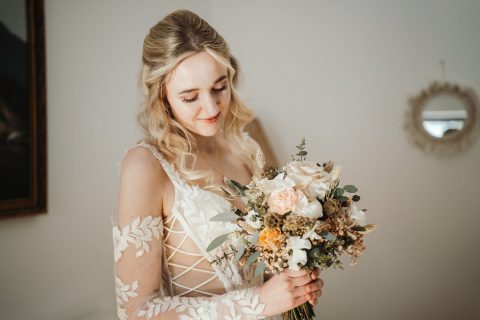 Hochzeitswahn - Eine Frau in einem weißen Spitzenbrautkleid hält einen Blumenstrauß und blickt nach unten. Sie steht in einem Innenbereich neben einer weißen Wand mit einem runden Spiegel – ein eleganter Moment, festgehalten von Nelyce Weddings, den Spezialisten für unvergessliche Hochzeitsplanung.
