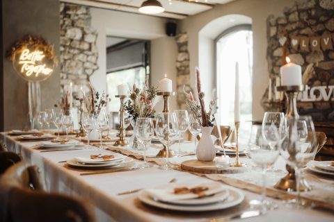 Hochzeitswahn - Ein langer Esstisch, gedeckt für eine formelle Veranstaltung, mit weißen Tellern, Gläsern, goldenen Kerzenleuchtern, Blumenarrangements und brennenden Kerzen. Im Hintergrund befinden sich Steinmauern und ein Neonschild mit der Aufschrift „Better Together“ – die perfekte Inspiration von Nelyce Weddings.