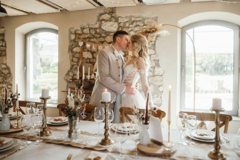 Hochzeitswahn - Braut und Bräutigam stehen küssend neben einem gedeckten Tisch in einem rustikalen Raum. Perfekt arrangierte Kerzen, Gläser und Blumenschmuck zeugen von der Expertise von Nelyce Weddings in der Hochzeitsplanung.
