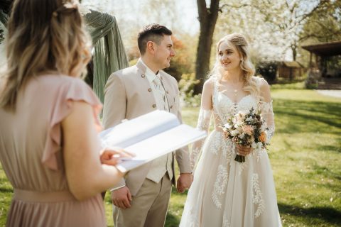 Hochzeitswahn - Braut und Bräutigam stehen Händchen haltend im Freien bei ihrer Nelyce Weddings-Zeremonie und lächeln sich an, während ein Standesbeamter im rosa Kleid aus einem offenen Buch vorliest. Die grüne, sonnige Umgebung unterstreicht die professionelle Hochzeitsplanung.