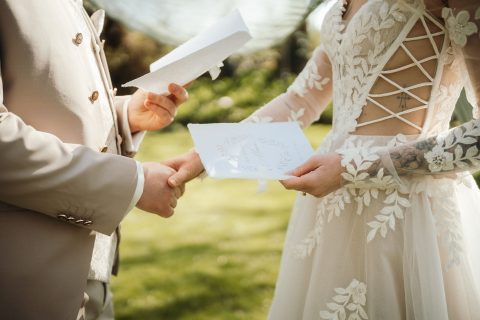 Hochzeitswahn - Ein Paar steht im Freien, hält Händchen und tauscht bei der Zeremonie von Nelyce Weddings das Eheversprechen aus. Die Braut trägt ein weißes Spitzenkleid mit floralen Details, der Bräutigam einen beigen Anzug – beide halten ihr Eheversprechen im Rahmen ihrer individuellen Hochzeitsplanung ab.