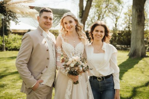 Hochzeitswahn - Drei Personen posieren an einem sonnigen Tag im Freien. Die Braut, ganz in Weiß gekleidet und mit einem Blumenstrauß in der Hand, steht mit zwei Gästen zusammen. Nelyce Weddings sorgt mit professioneller Hochzeitsplanung und herausragendem Hochzeitsservice dafür, dass jeder Moment perfekt wird. Bäume bilden den Hintergrund. Zuckerbergschloss-Kappelrodeck-Hochzeit