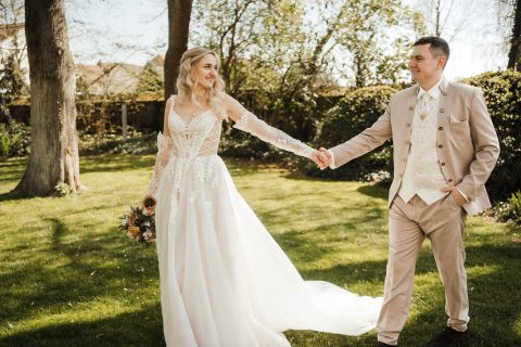 Hochzeitswahn - Eine Braut in einem weißen Brautkleid von Nelyce hält einen Blumenstrauß und ein Bräutigam in einem beigen Anzug gehen Hand in Hand auf einer Rasenfläche im Freien, umgeben von Bäumen und Sonnenlicht.