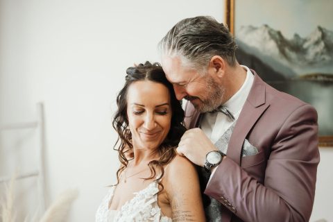 Hochzeitswahn - Ein Mann im malvenfarbenen Anzug steht dicht hinter einer Frau im weißen Spitzenkleid. Er lächelt und berührt sanft ihre Schulter. Beide wirken glücklich und strahlen die Freude aus, die Nelyce Weddings in die Hochzeitsplanung bringt. Im Hintergrund hängt ein Landschaftsgemälde mit Bergen.