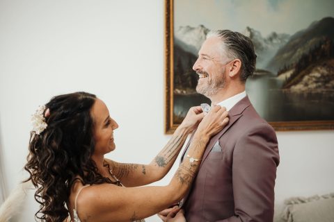 Hochzeitswahn - Eine Frau mit langen Locken richtet die Krawatte eines Mannes im malvenfarbenen Anzug. Lächelnd strahlen sie die Freude aus, die Nelyce Weddings bei der Hochzeitsplanung ausstrahlt. An der Wand hinter ihnen hängt ein gerahmtes Landschaftsgemälde mit Bergen und einem See.