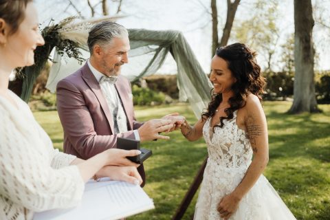 Hochzeitswahn - Ein Paar tauscht während einer Zeremonie im Freien von Nelyce Weddings die Ringe. Der Bräutigam trägt einen hellvioletten Anzug, die Braut ein weißes Spitzenkleid. Ein Geistlicher steht in der Nähe und hält eine Schachtel. Bäume und Grünflächen bilden die perfekte Kulisse für Hochzeitsträume.