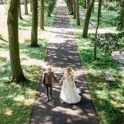 Hochzeitswahn - An einem sonnigen Tag gehen Braut und Bräutigam Hand in Hand einen von Bäumen gesäumten Weg in einem Park entlang. Eingefangen von Jens Schaffhirt Photography, erzeugen die Schatten der Bäume beim Spazierengehen kunstvolle Muster auf dem Weg.