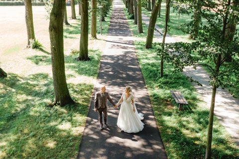 Hochzeitswahn - An einem sonnigen Tag gehen Braut und Bräutigam Hand in Hand einen von Bäumen gesäumten Weg in einem Park entlang. Eingefangen von Jens Schaffhirt Photography, erzeugen die Schatten der Bäume beim Spazierengehen kunstvolle Muster auf dem Weg.