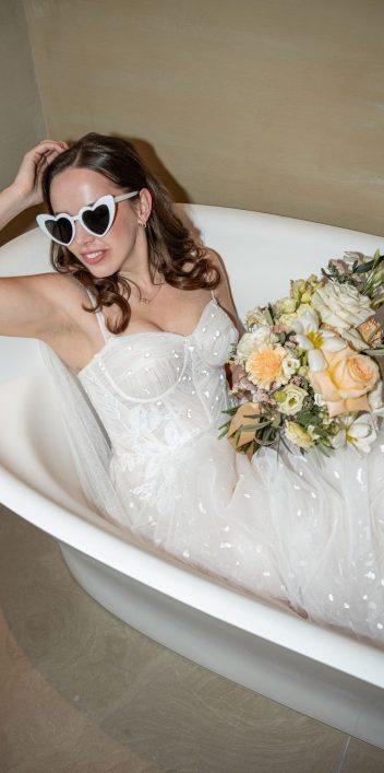 Hochzeitswahn - Eine Braut in einem weißen trägerlosen Brautkleid und einer herzförmigen Sonnenbrille sitzt in einer weißen Badewanne und hält einen Strauß gelber und weißer Blumen in der Hand – ein automatisch gespeicherter Entwurf aus Eleganz und Verspieltheit. PURS-Hotel-Hochzeit