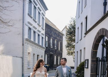 Hochzeitswahn - Eine Braut in einem trägerlosen Brautkleid und ein Bräutigam in einem hellgrauen Anzug gehen Hand in Hand eine mit Kopfsteinpflaster gepflasterte Straße entlang, die von weißen Gebäuden gesäumt ist. Beide lächeln und schauen sich an – ein Automatisch gespeicherter Entwurf puren Glücks. PURS-Hotel-Hochzeit