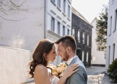 Hochzeitswahn - Braut und Bräutigam stehen eng beieinander auf einer gepflasterten Straße, umarmen sich und schauen sich in die Augen. Das Sonnenlicht beleuchtet ihre Gesichter, und dieser Moment fühlt sich an wie ein automatisch gespeicherter Entwurf – Liebe in ihrer reinsten Form. PURS-Hotel-Hochzeit