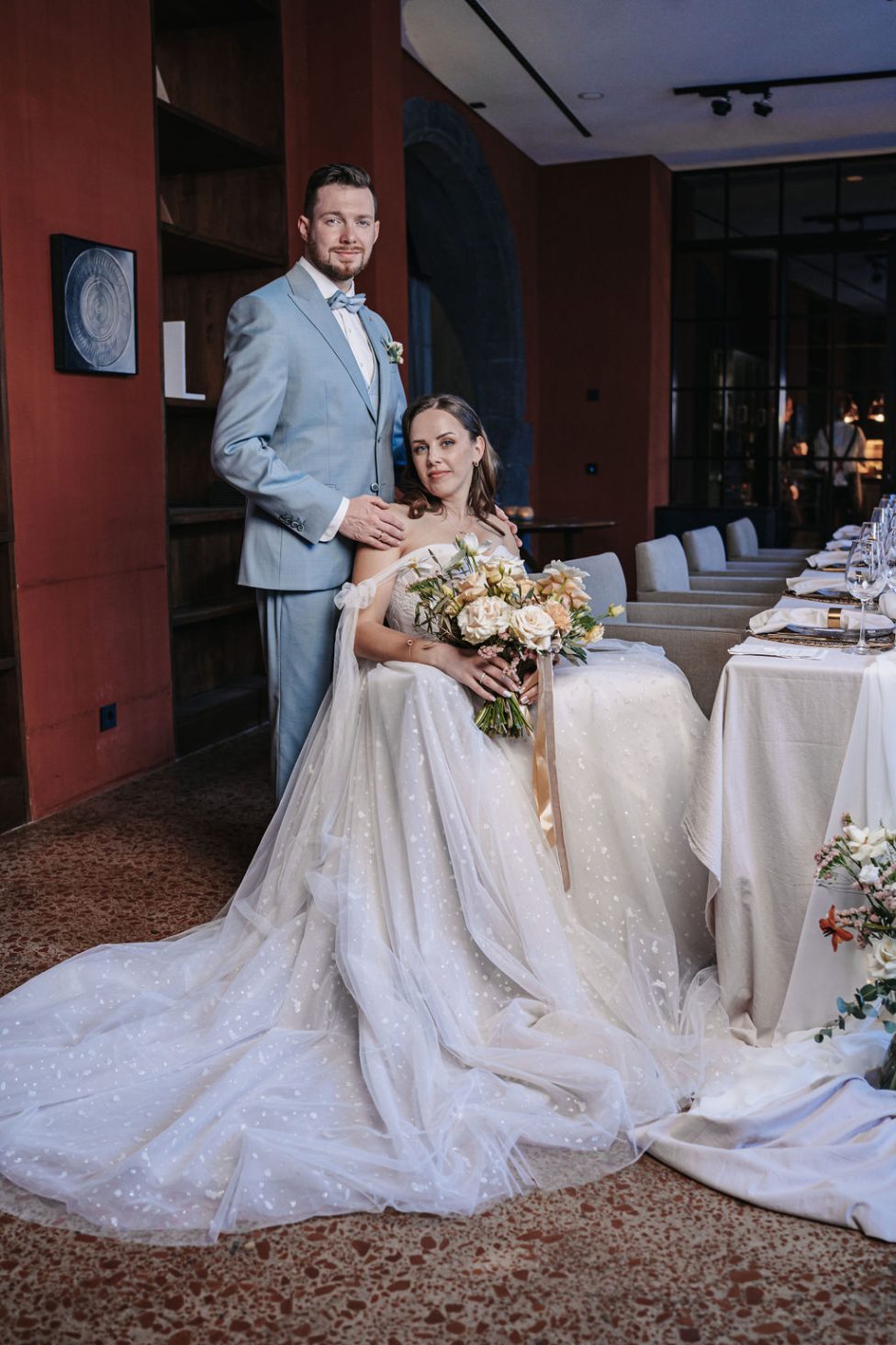 Hochzeitswahn - Eine Braut in einem weißen schulterfreien Brautkleid mit langer Schleppe sitzt mit einem Blumenstrauß in der Hand da, während ein Bräutigam in einem hellblauen Anzug neben ihr steht und seine Hand auf ihrer Schulter hat – eine automatisch elegante Szene wie ein automatisch gespeicherter Entwurf. PURS-Hotel-Hochzeit