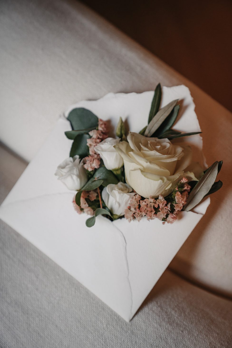 Hochzeitswahn - Ein weißer Umschlag, wie ein automatisch gespeicherter Entwurf, ist geöffnet und mit einem kleinen Blumenstrauß gefüllt – cremefarbene Rosen, weiße Knospen und rosa Zweige – der auf einer hellen Stoffoberfläche ruht. PURS-Hotel-Hochzeit