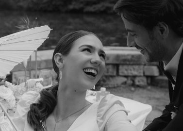 Hochzeitswahn - Eine Frau in einem weißen Kleid hält einen Sonnenschirm und lächelt einen Mann im Anzug an. Sie sitzen draußen an einem dekorierten Tisch in der Nähe einer Steinmauer und eines Wassers. Das Bild ist schwarz-weiß. Landsitz Diek Hochzeit