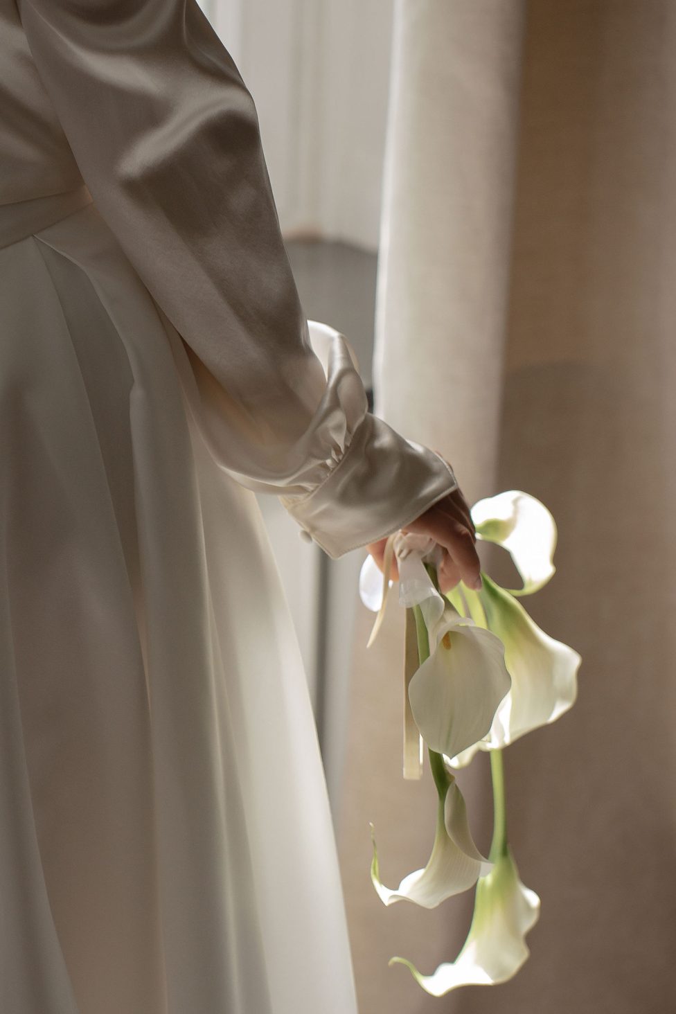 Hochzeitswahn - Eine Person in einem langärmeligen, satinartigen weißen Kleid hält einen kleinen Strauß weißer Callas an den Stielen und steht neben einem Fenster mit sanftem Tageslicht. Landsitz Diek Hochzeit