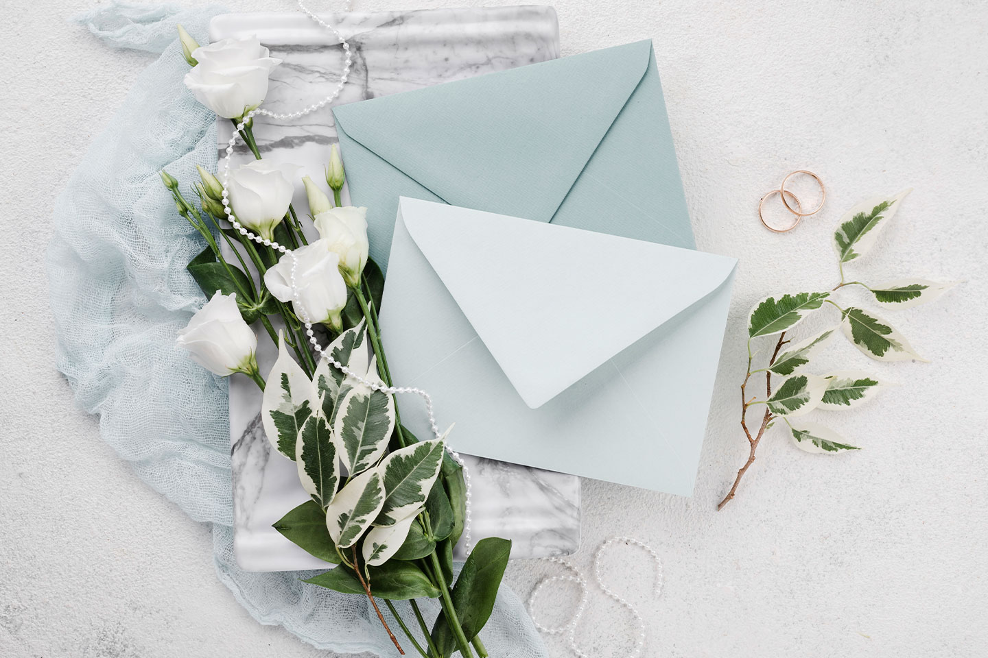 Hochzeitswahn - Zwei blaue Umschläge, weiße Blumen, Blätter, zwei goldene Ringe und eine Perlenkette auf Marmor und hellblauem Stoff - stilvolle visuelle Elemente für eine elegante Hochzeitseinladung auf weißer, strukturierter Oberfläche arrangiert. Landsitz Diek Hochzeit
