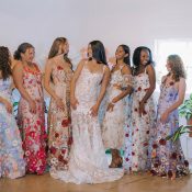 Hochzeitswahn - Eine Gruppe von sieben Brautjungfern in geblümten Kleidern steht in einem Innenraum in einem Halbkreis. Die Frau in der Mitte trägt ein weißes geblümtes Kleid, und alle lächeln. An den Wänden und Fenstern sind Topfpflanzen zu sehen.